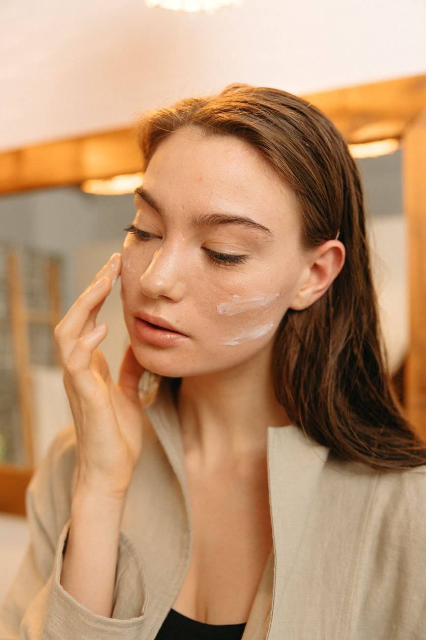 A woman gently applies skincare cream on her face indoors with natural light.